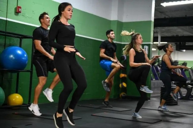 Group of five people exercising with high knees in a gym with green and white walls and fitness equipment.