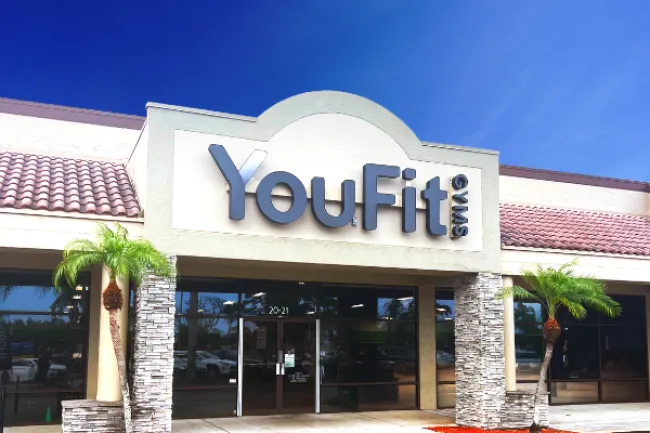 YouFit Gyms storefront with signage, palm trees, and blue sky in a shopping plaza setting.