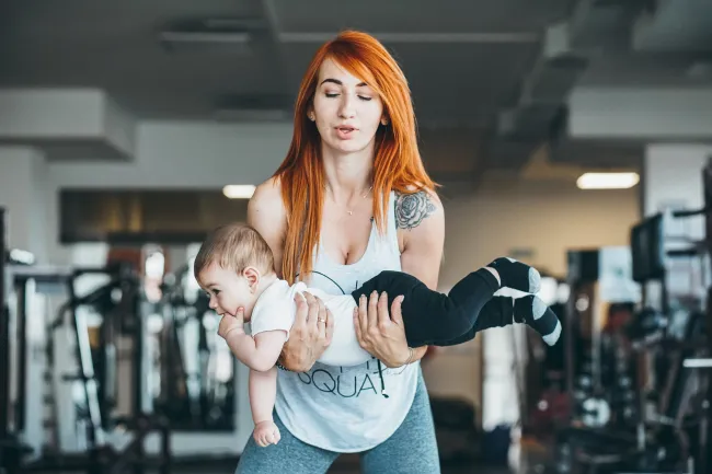 Young woman with red hair exercising by lifting a baby in a gym setting with workout equipment in the background