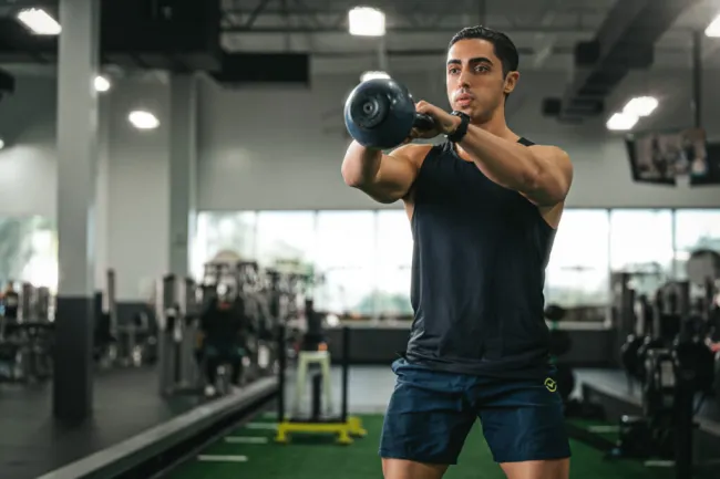Man in black tank top and shorts lifting a kettlebell during a workout in a gym with exercise equipment.