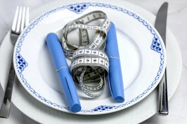 Blue insulin pens and a white measuring tape on a white plate with blue patterns, fork and knife beside.