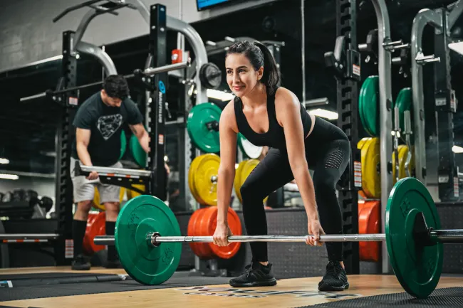 Woman lifting barbell with weights in gym while a man adjusts equipment in the background
