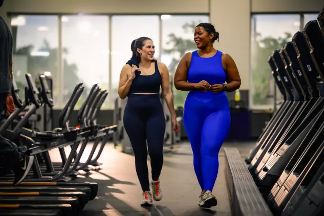Two women in activewear walking and smiling in a gym surrounded by cardio equipment and natural light.