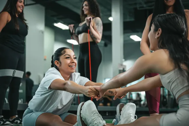 Group of women exercising together in a gym, smiling and stretching with workout bands and holding hands.
