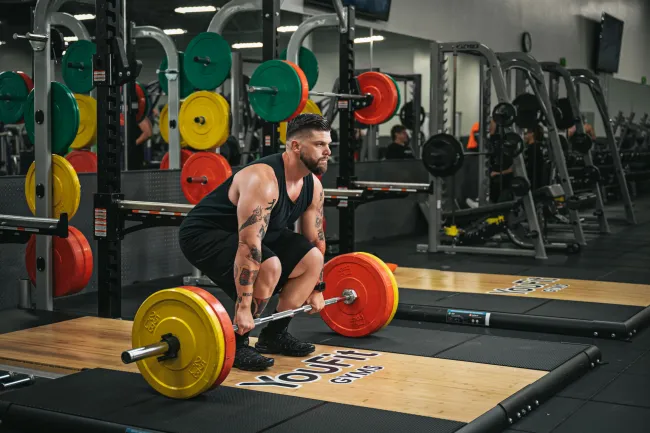Man with tattoos preparing to deadlift barbell with colorful weight plates in a modern gym.