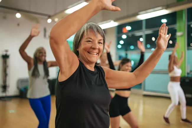 Smiling mature woman in black top leading group exercise class indoors with participants raising arms and moving.