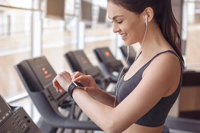 Smiling woman in gym adjusting smartwatch while wearing earphones near treadmills with large windows.