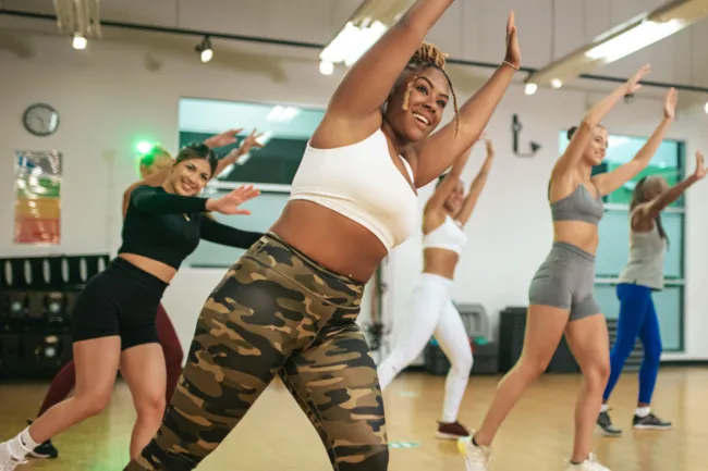 A group of five women engage in a lively dance class in a studio. They are smiling and wearing athletic wear, performing synchronized dance moves. The studio has wooden floors, mirrors, and fitness equipment in the background.