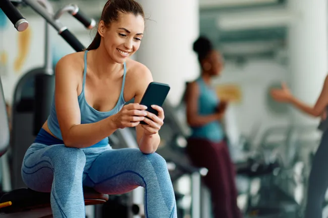 Smiling woman in blue workout clothes sitting on gym bench using smartphone with exercise equipment in background