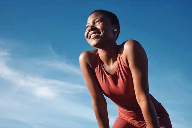 Smiling woman in red workout clothes wearing wireless earbuds outdoors under clear blue sky.