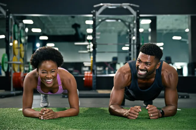 Smiling young man and woman doing planks together on artificial grass in a gym setting.