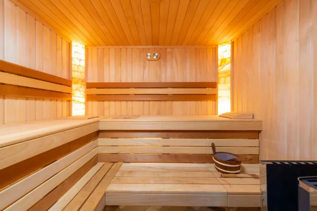 Interior of a wooden sauna with benches, backrests, warm lighting, and a sauna bucket with ladle.