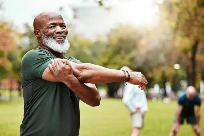 Smiling elderly man stretching his arm in a park with people exercising in the background on a sunny day