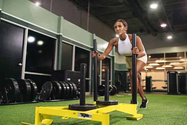 Woman in white athletic wear pushing a weighted sled on green turf in a gym setting.