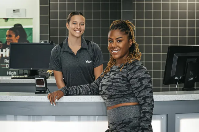 Smiling female fitness trainer and gym member at the reception desk in a modern gym setting.