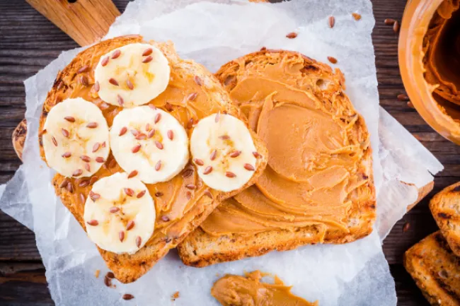 Peanut butter spread on toasted bread slices topped with banana slices and flax seeds on parchment paper.