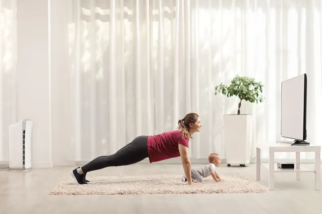 Mother and baby doing plank exercise together on a rug in a bright living room with TV and plant.