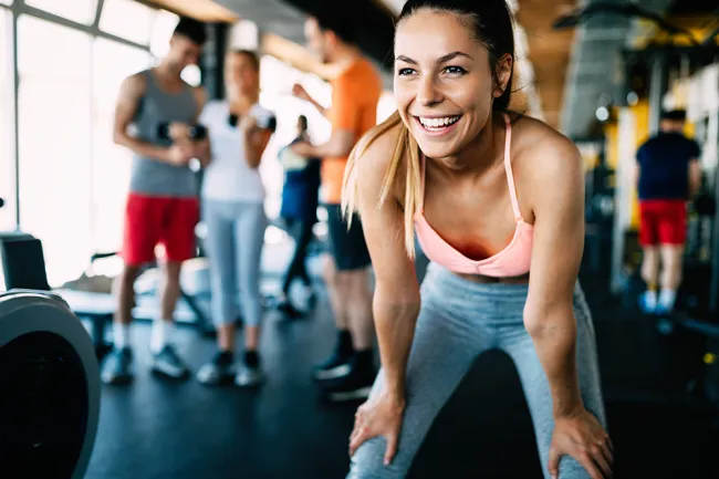 Smiling woman in pink sports bra leaning forward in gym with people training in the background