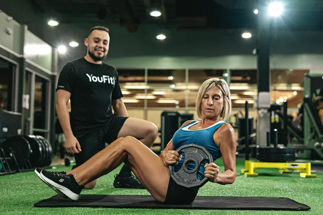 Woman performing Russian twists with weight plate guided by male personal trainer in gym setting.