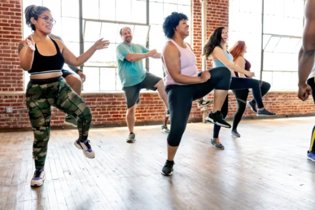 Group of diverse adults exercising and stretching together in a bright fitness studio with large windows.