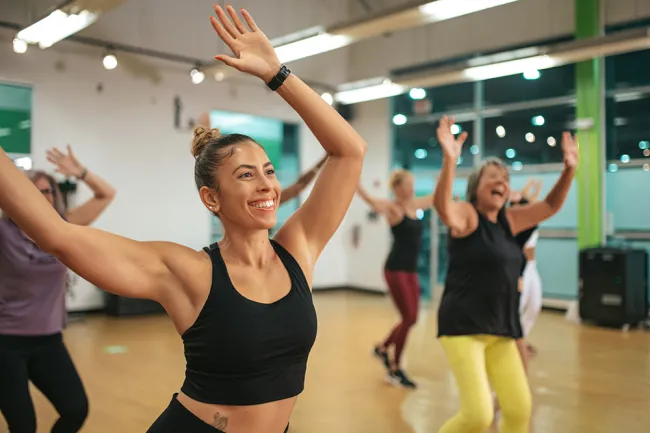 Women of different ages happily participating in a group fitness class with raised arms indoors.