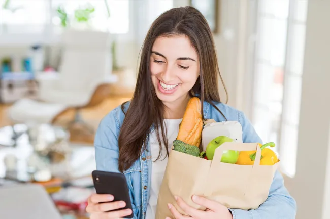 Smiling young woman holding groceries and checking phone in bright modern kitchen at home