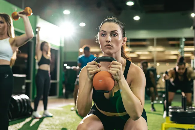 Focused woman exercises with kettlebell in gym surrounded by others working out with dumbbells and weights.
