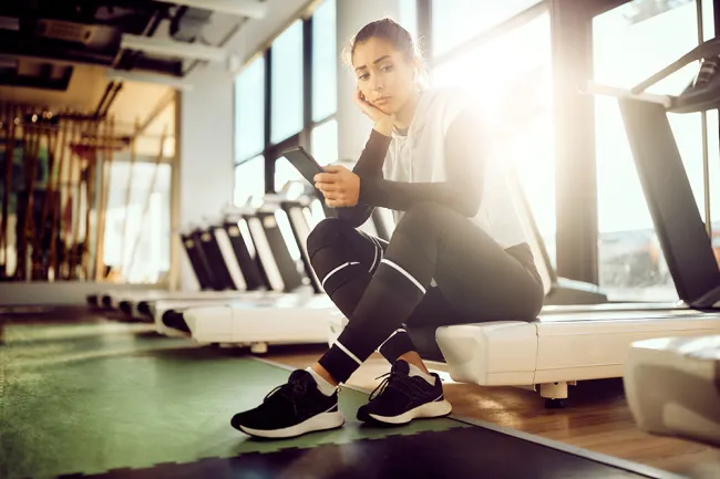 Young woman in workout clothes sitting on treadmill in gym looking at phone with thoughtful expression.