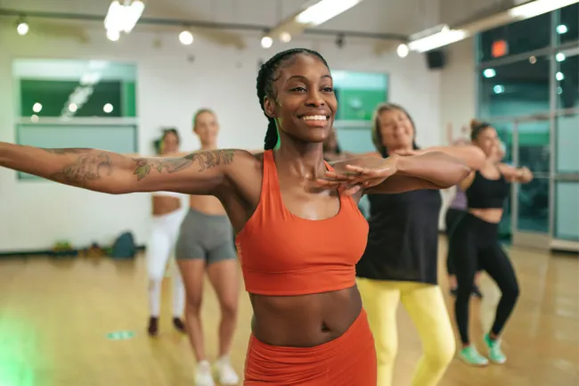 Smiling woman in an orange sports outfit leading a diverse group in an exercise class in a studio.