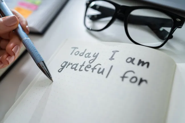 Person writing gratefulness journal entry with glasses and notebook nearby on white desk.