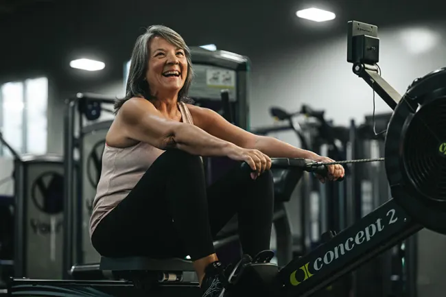 Mature woman smiling while using a rowing machine for exercise in a modern gym setting.