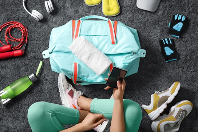 Person in athletic wear sitting on floor surrounded by gym bag, sneakers, water bottle, gloves, and headphones.