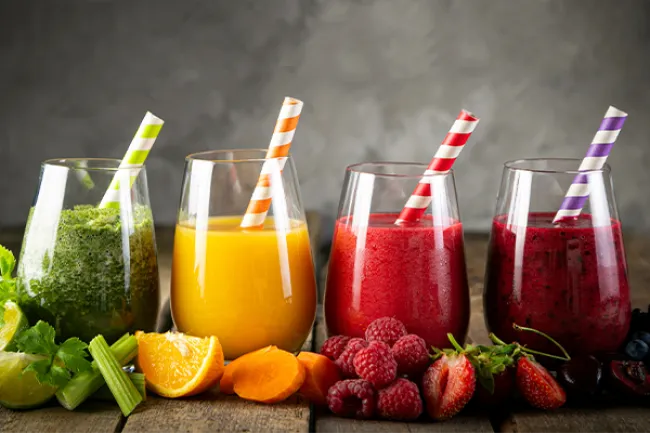 Four colorful fruit and vegetable smoothies with striped straws and fresh produce on a wooden surface.