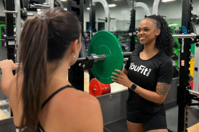 Personal trainer assisting woman with weightlifting at gym with green weight plate and fitness equipment.