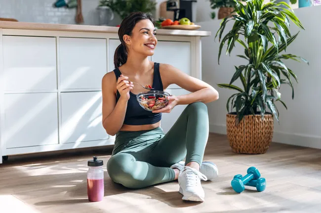 Woman in workout clothes sitting on floor eating a bowl of fruit, with dumbbells and a smoothie nearby in bright kitchen.
