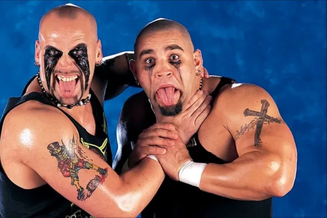 Two muscular men with face paint and tattoos pose playfully, one choking the other who sticks out his tongue.
