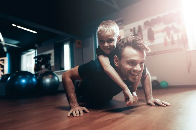 Man doing push-ups with a young boy on his back in a bright gym setting, both smiling and having fun.