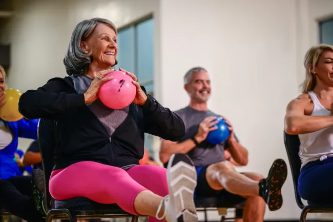 Seniors participating in a seated fitness class holding colorful exercise balls and stretching their legs.