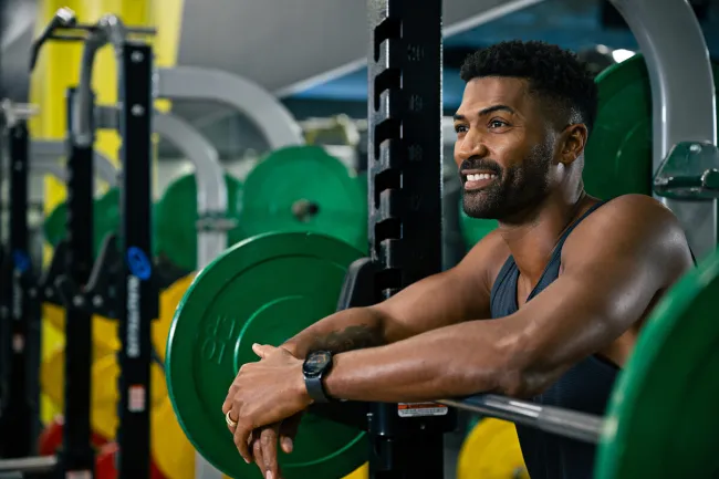 Smiling man resting on barbell loaded with green weight plates in a gym setting.