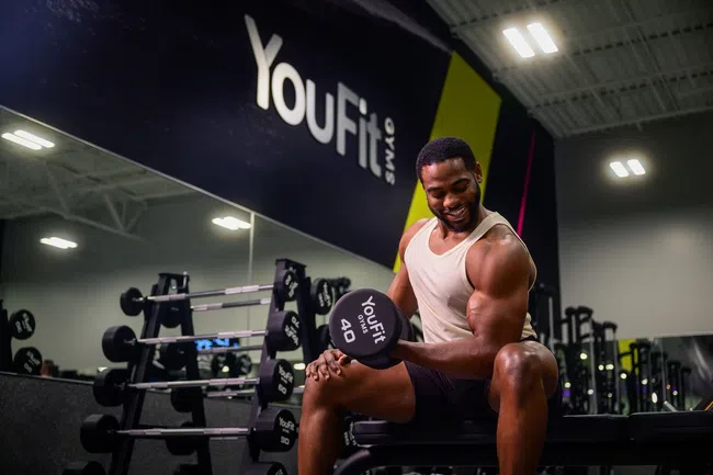 Smiling man lifting 40-pound dumbbell in YouFit gym with workout equipment and mirror in the background