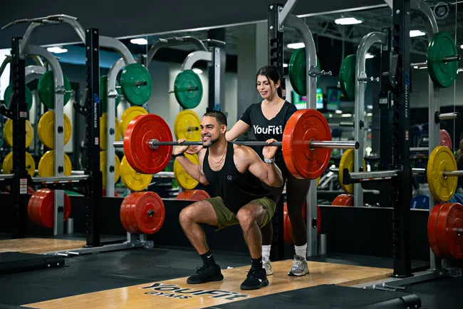 Man performs barbell squat with a trainer spotting him in a gym surrounded by colorful weight plates and mirrors.