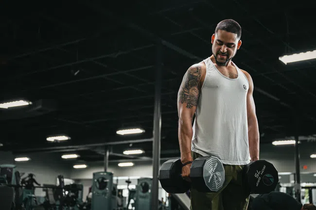 Man lifting dumbbells in YouFit Gyms wearing a white tank top and green pants under bright lights