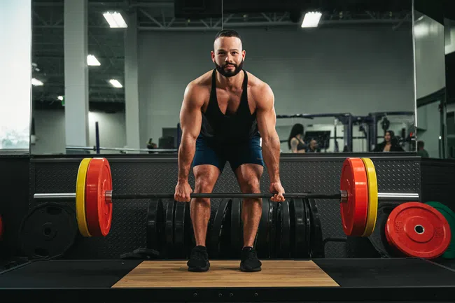Muscular man wearing black tank top lifting heavy barbell with colorful weight plates in gym.