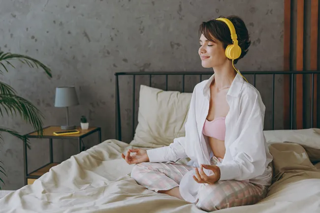 Young woman in pajamas meditating on bed with yellow headphones in cozy bedroom with soft lighting.