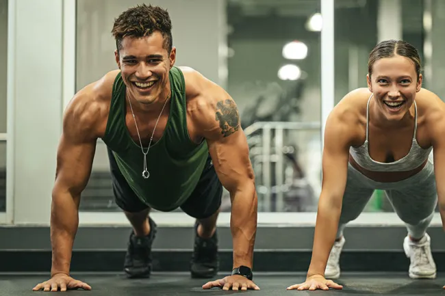 Smiling man and woman doing push-ups together in a gym with weights and mirrors in background