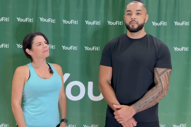 Fitness trainer and participant posing in front of YouFit gym backdrop wearing workout attire.