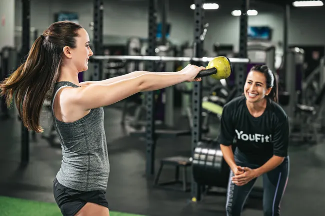 Woman lifting a kettlebell while a trainer encourages her in a gym setting with fitness equipment.