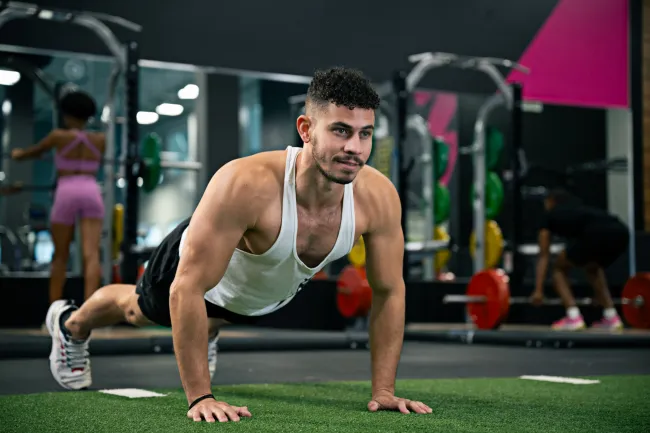 Man in white tank top doing push-ups on gym turf with weightlifting equipment and people in background