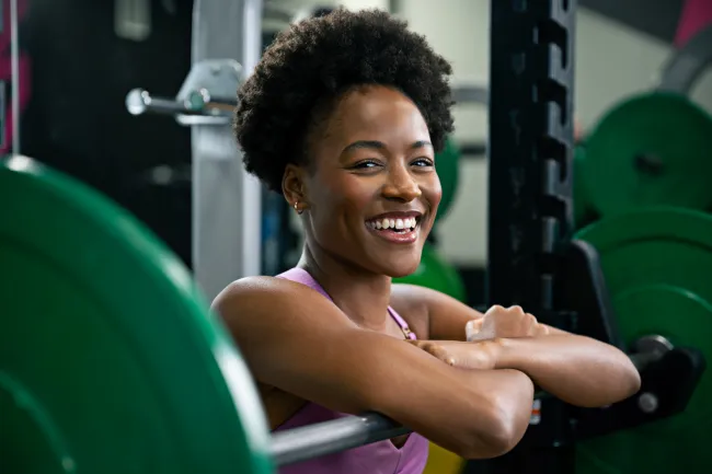 Smiling young woman resting on a barbell in a gym with weight plates in the background, ready for weightlifting.