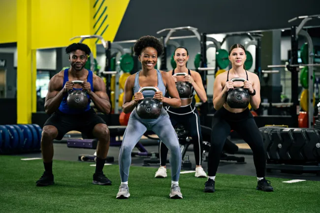 Four diverse adults performing kettlebell squats together in a bright, modern gym with weight equipment in background
